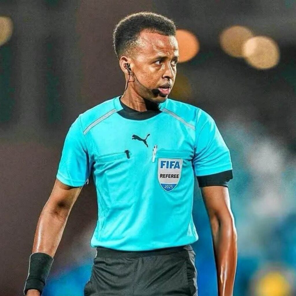 Árbitro de futebol em uniforme teal caminha em campo iluminado, usando fone de ouvido e microfone.