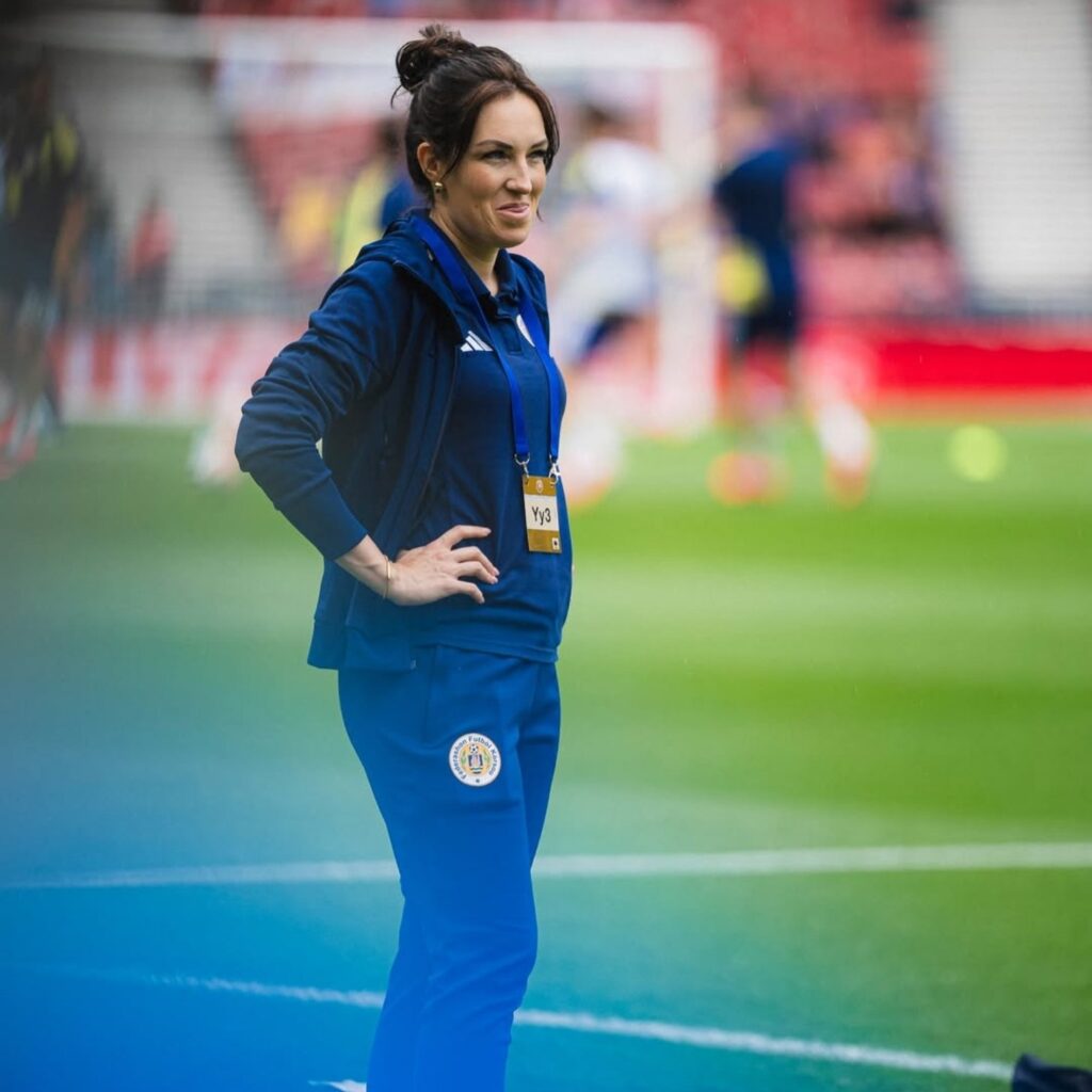 Mulher em agasalho azul da Adidas observa o campo de futebol com as mãos na cintura.
