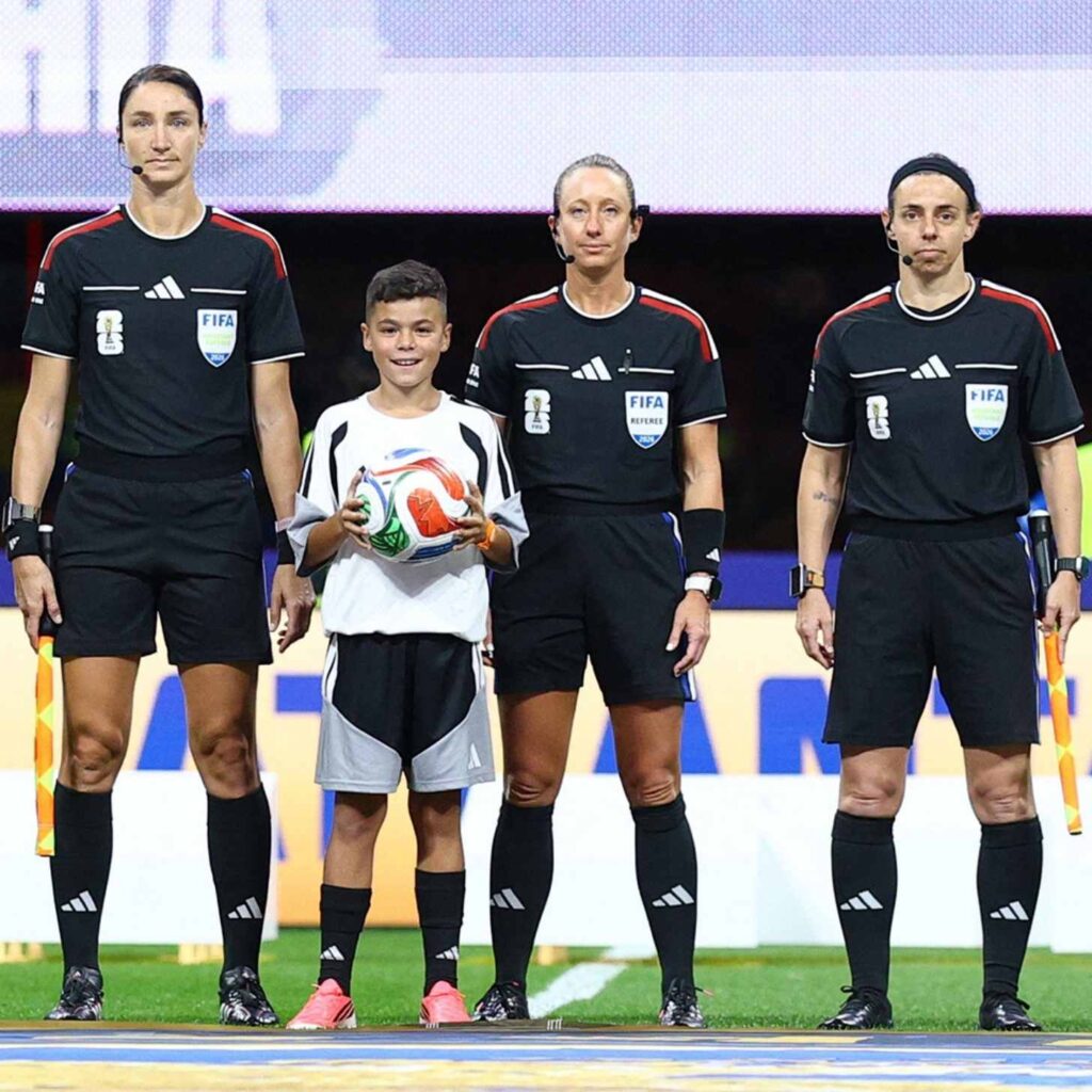 Três árbitras em uniformes da FIFA ao lado de um jovem mascote segurando a bola do jogo em campo.