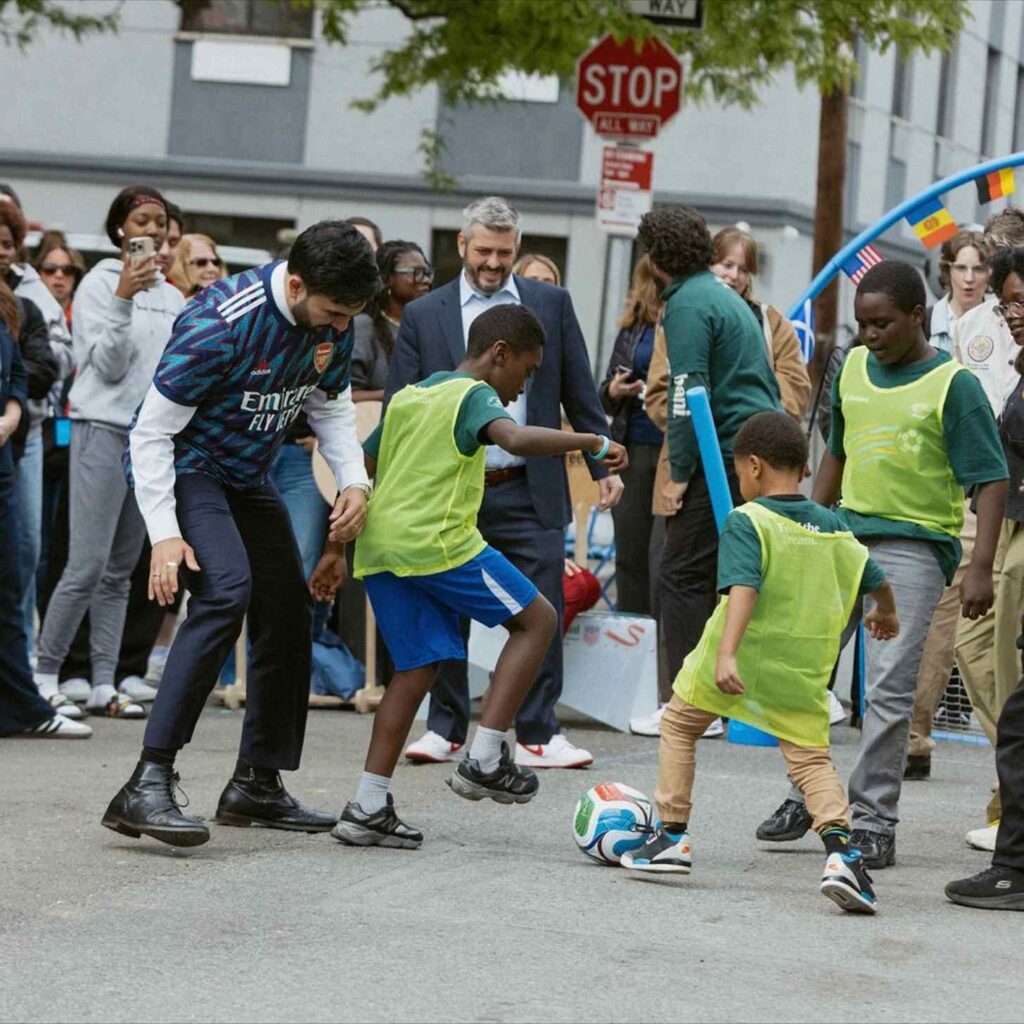 Homem joga futebol de rua com crianças em coletes amarelos, enquanto uma multidão assiste em Boston.